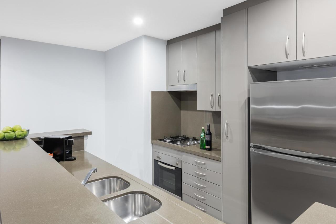 Meriton Suites Kent Street, Sydney Exterior foto The photo depicts a modern kitchen featuring a compact design. It includes gray cabinetry with several drawers and cabinets, a countertop with two sinks, and stainless steel appliances. There is a stove and oven integrated into the cabinetry, along w