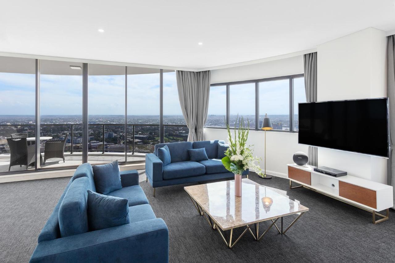 Meriton Suites Kent Street, Sydney Exterior foto The photo shows a modern living room with large windows that provide a panoramic view of the city. The room features two blue sofas arranged around a stylish marble-top coffee table. There are decorative items, including a vase with flowers on the ta